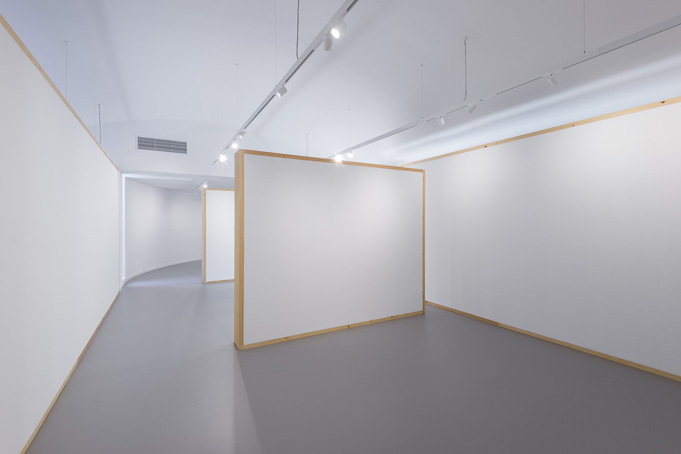 Interior view featuring modular white walls with wooden frames, a curved passageway in the background, and overhead track lighting.