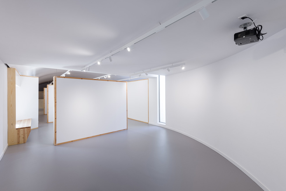 Curved gallery space with modular white walls and wooden frames, a bench built into the wall, overhead track lighting, and a ceiling-mounted projector.