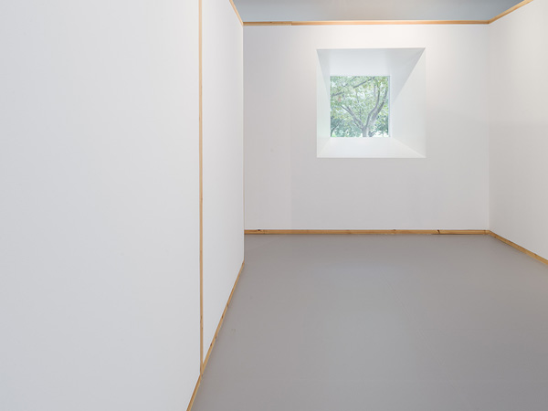 Minimalistic interior with white walls and wooden frames, featuring a single window providing a view of trees outside.