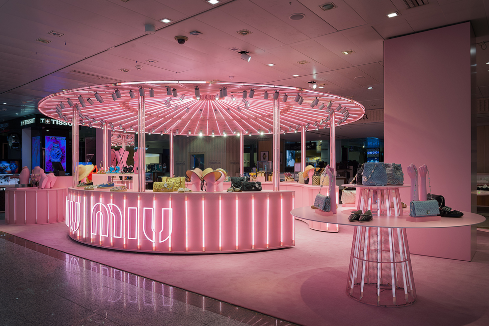 Pink-themed Miu Miu pop-up store with a circular display area, illuminated by vibrant pink neon lights, showcasing various handbags and accessories.