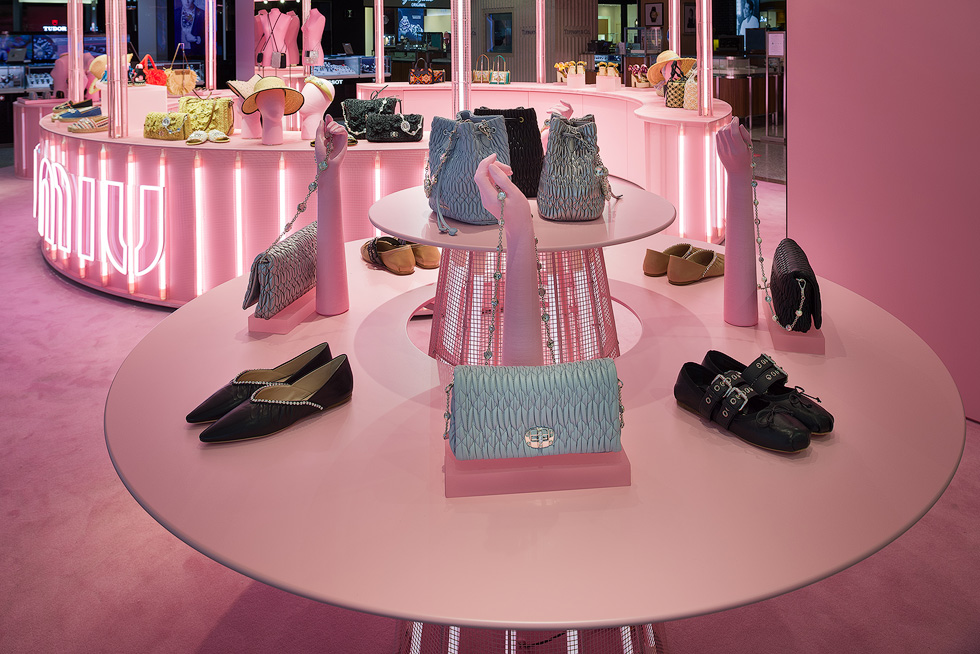 Close-up of a pink-themed Miu Miu pop-up store display, featuring handbags and shoes arranged on a two-tier round table with a conic base illuminated from within by neon lighting. Standing on the table, three pink mannequin hands showcasing accessories.