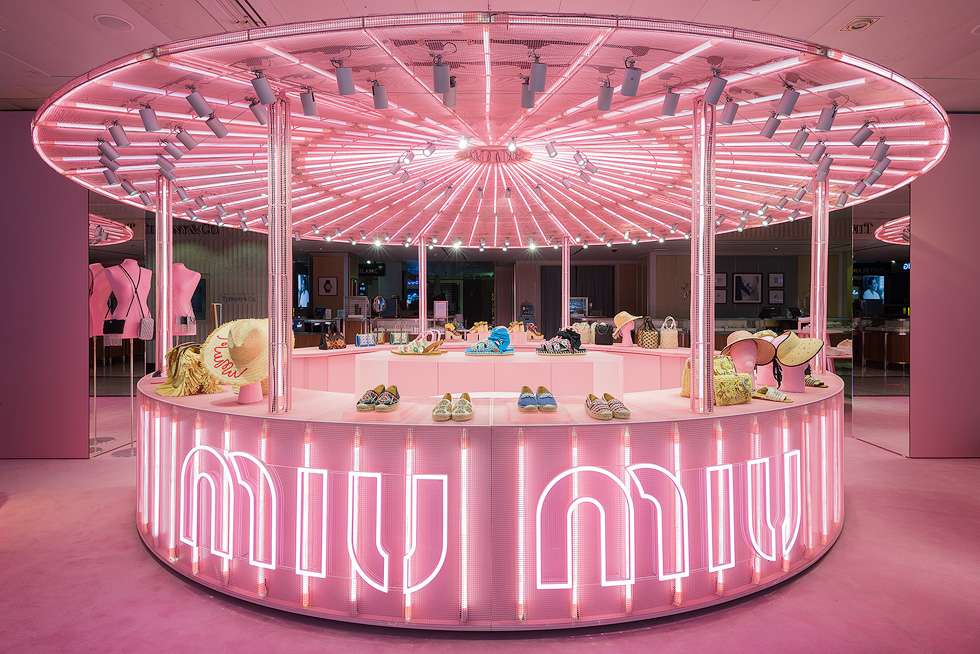 Illuminated pink Miu Miu pop-up store display with handbags, shoes, and hats under a canopy of bright pink lights.