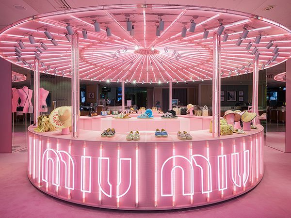 Illuminated pink Miu Miu pop-up store display with handbags, shoes, and hats under a canopy of bright pink lights.