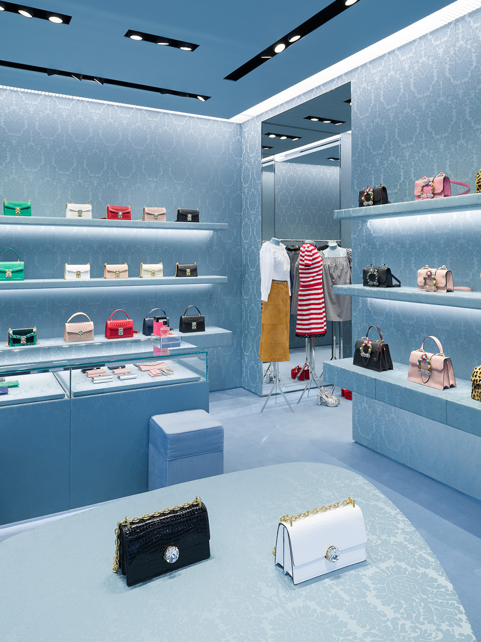 A stylish Miu Miu store interior, with elegant display shelves featuring a variety of colorful handbags, glass counters showcasing smaller accessories, and on the foreground, a black bag side by side a white bag on a blue patterned oval table.