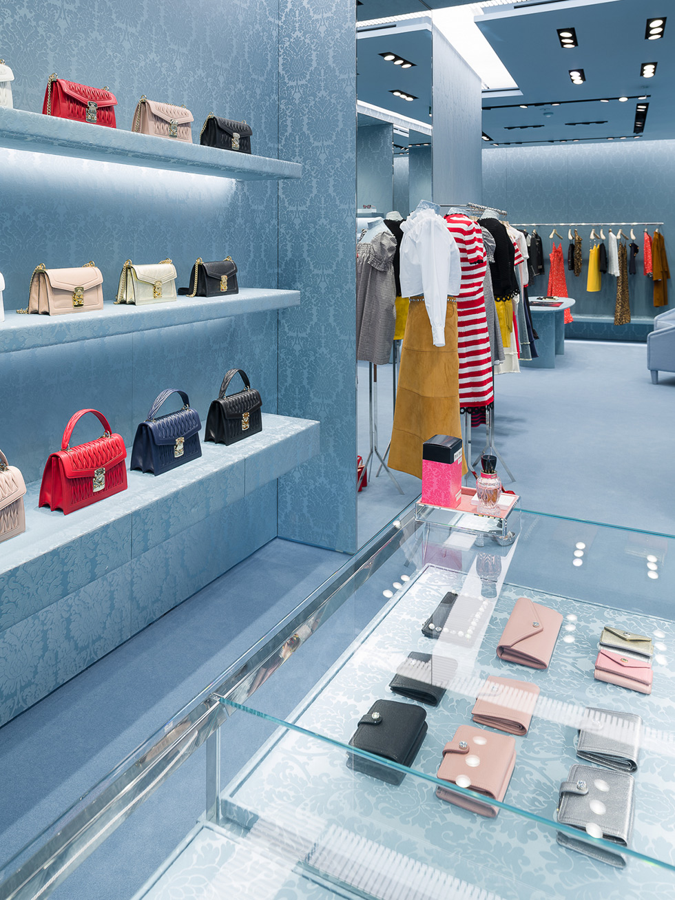 Miu Miu store interior featuring shelves with handbags, a glass display counter with wallets, and clothing racks with various garments.