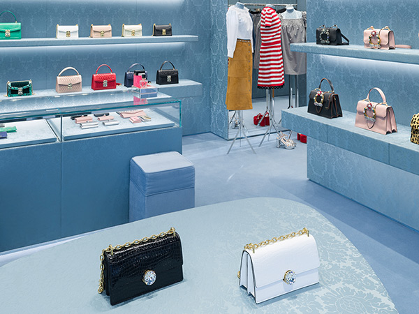 A stylish Miu Miu store interior, with elegant display shelves featuring a variety of colorful handbags, glass counters showcasing smaller accessories, and on the foreground, a black bag side by side a white bag on a blue patterned oval table.