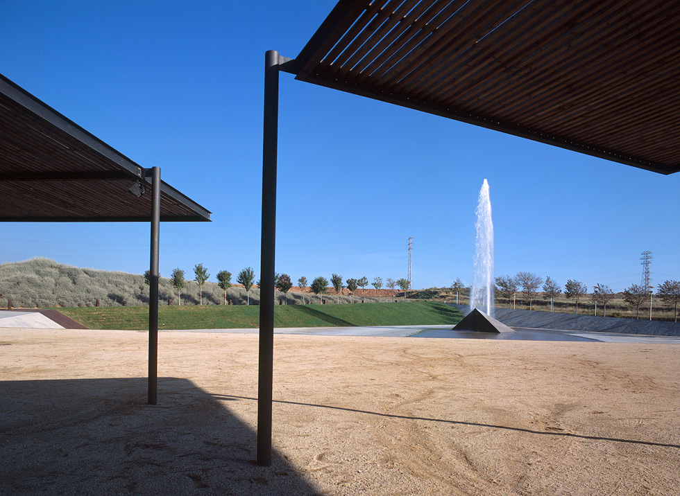 Outdoor space with two steel and wood modern shade structures and a large central jet water fountain against a clear blue sky.