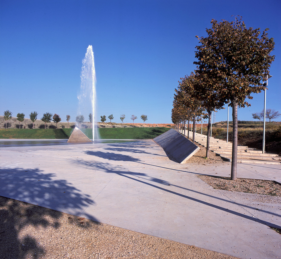 A line of trees along a walkway with gentle steps besides a large jet water fountain in an open park area.