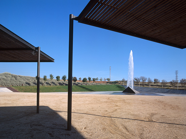 Outdoor space with two steel and wood modern shade structures and a large central jet water fountain against a clear blue sky.