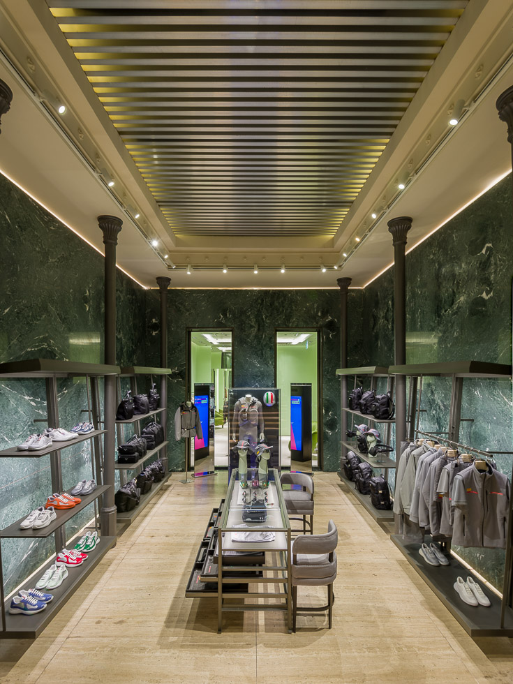 Luxurious boutique interior with green marble walls, elegant lighting, and black metal shelving displaying sneakers, bags, and gray apparel. A central glass table showcases accessories, while a doorway at the back leads to another section.