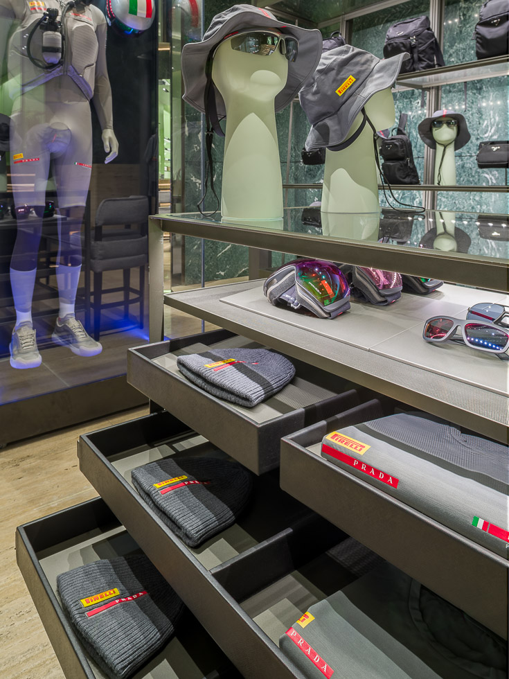 Luxury Prada boutique, a glass table with half opened metal drawers showcasing gray apparel displaying the brands Prada and Pirelli. In the background a mannequin in a performance outfit is encased in a glass display.