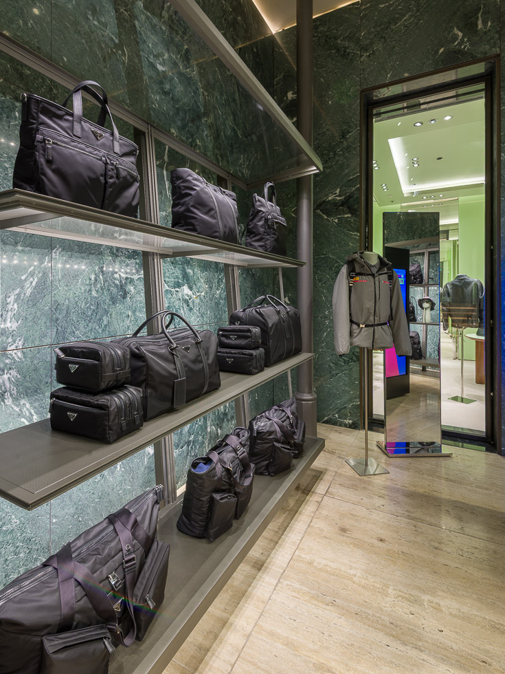 Luxury Prada boutique with green marble walls, showcasing designer black bags on sleek metal shelving. A mannequin with gray apparel and a backpack besides a mirror and an opening onto another section.