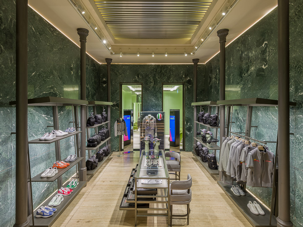 Luxurious boutique interior with green marble walls, elegant lighting, and black metal shelving displaying sneakers, bags, and gray apparel. A central glass table showcases accessories, while a doorway at the back leads to another section.