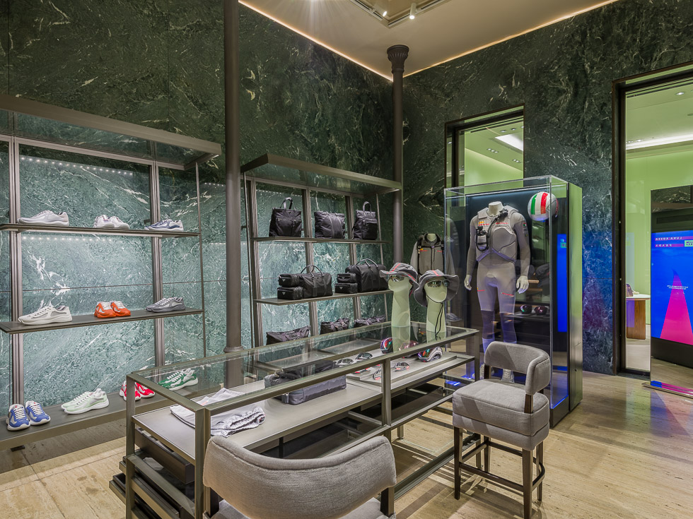 High-end boutique interior with green marble walls, sleek metal shelving showcasing sneakers and bags, and a glass display case featuring a mannequin in a performance outfit with a racing helmet. A central glass table holds accessories, with plush seating nearby.