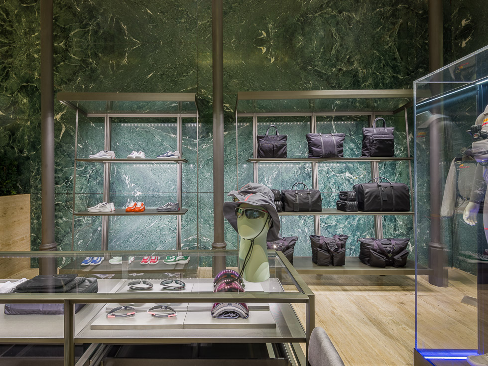 Luxury boutique with green marble walls, showcasing designer sneakers, black bags, and accessories on sleek metal shelving. A glass table displays eyewear and hats, while a mannequin in a performance outfit is encased in a glass display.