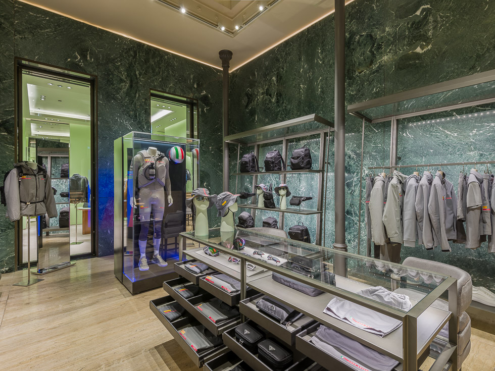 Luxury Prada boutique with green marble walls, showcasing designer sneakers, black bags, and accessories on sleek metal shelving. A glass table displays eyewear and hats, while a mannequin in a performance outfit is encased in a glass display.