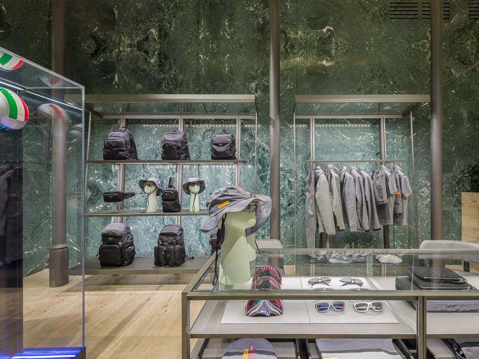 Luxury boutique with green marble walls, showcasing designer sneakers, black bags, and accessories on sleek metal shelving. A glass table displays eyewear and hats, while a mannequin in a performance outfit is encased in a glass display.