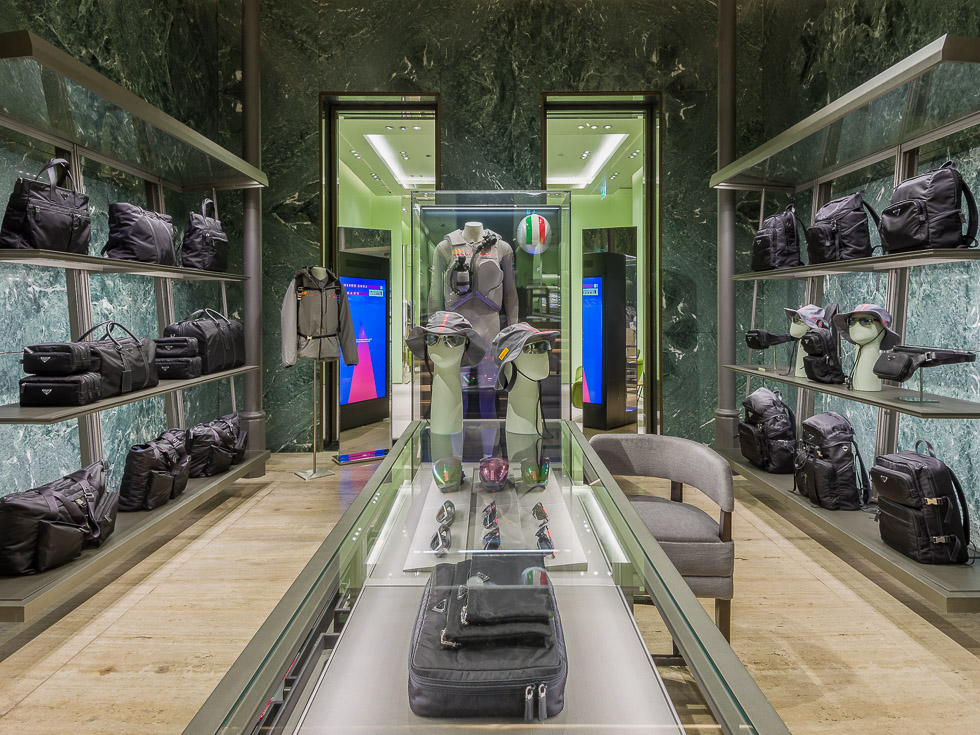 Luxury Prada boutique with green marble walls, showcasing designer sneakers, black bags, and accessories on sleek metal shelving. A glass table displays eyewear and hats, while a mannequin in a performance outfit is encased in a glass display.