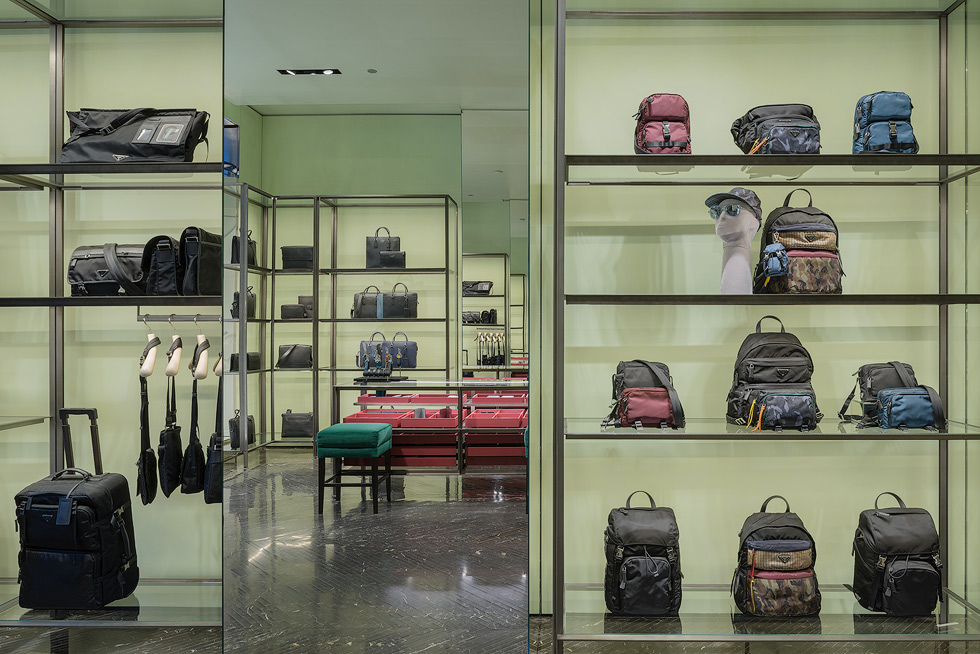 Luxury store interior with mirrors showcasing various designer bags and backpacks displayed on sleek metal shelves against a pale green backdrop.