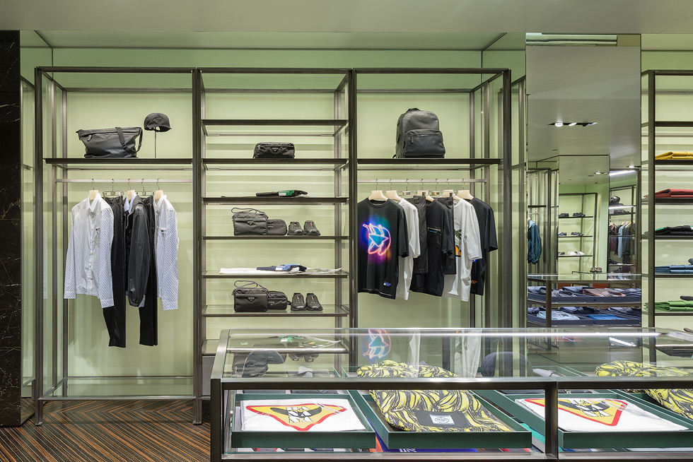 Luxury retail store interior with neatly arranged shelves displaying designer bags, clothing, and accessories against a Prada green backdrop.