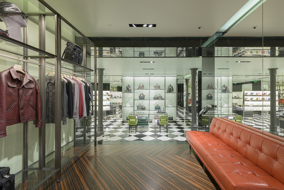 Prada store interior with clothing racks, leather jackets, and a checkered floor, featuring a red sofa and display shelves in the background.