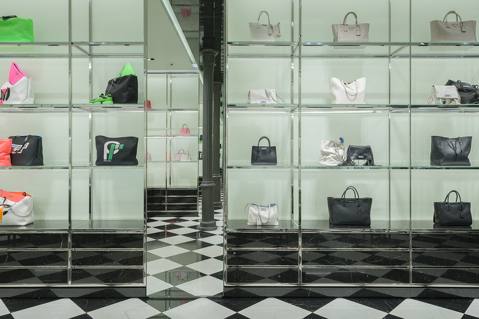 Shelves of high-end handbags in various colors displayed against a light green background in a Prada store, with two facing mirrors and a black cast iron column caught in the infinity mirror effect.