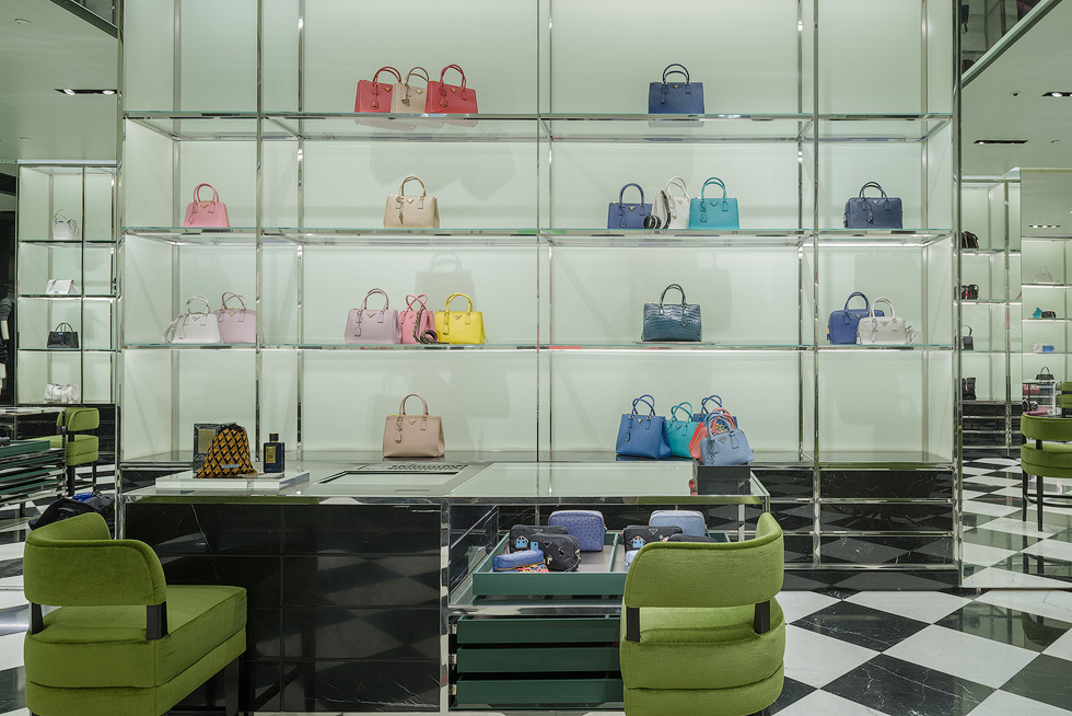 Elegant display of luxury handbags in a Prada store, with colorful bags arranged on glass shelves and green chairs in the foreground.