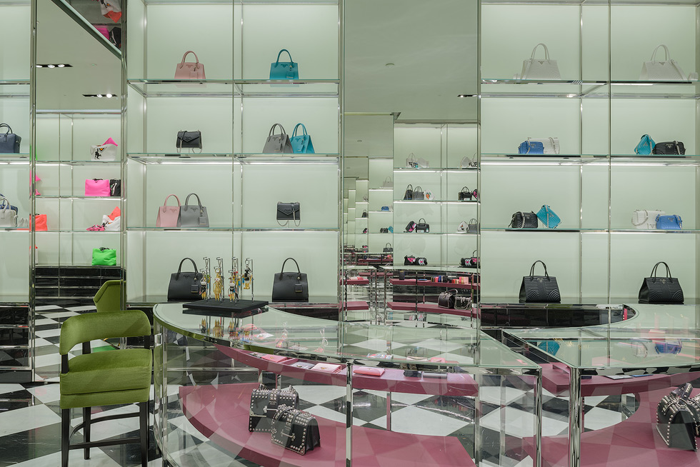 Elegant luxury store showcasing handbags on glass shelves, with a green chair and mirrored walls enhancing the display and featuring an infinity mirror effect.