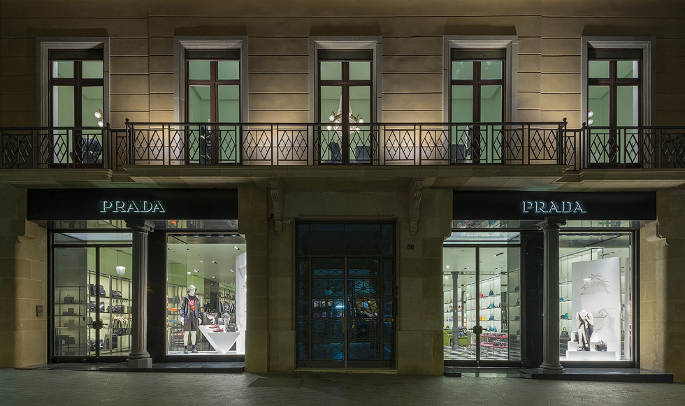 Exterior of a Prada store at night, showcasing brightly lit window displays with luxury items and mannequins, framed by a classic building facade.