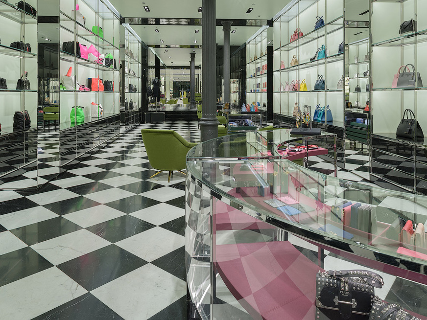 Prada store interior with black and white checkered flooring, glass shelves displaying colorful handbags, green chairs and black cast iron columns.