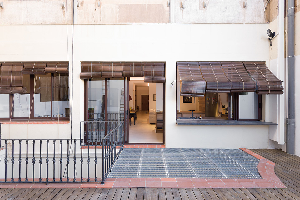 Outdoor patio with metal railing, wooden deck, and large windows with bamboo shades, leading into a modern interior.