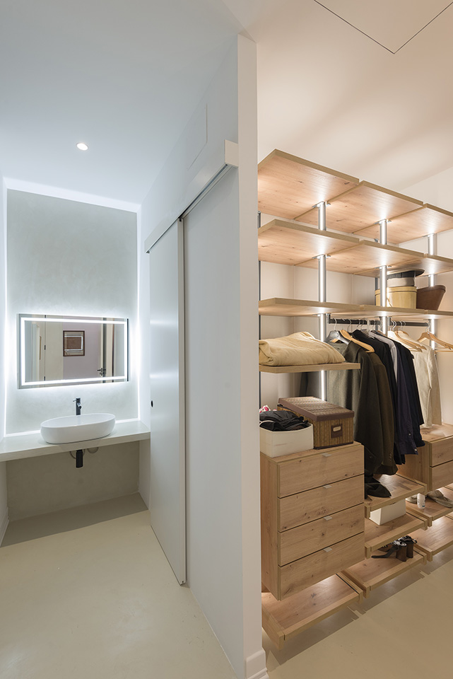 Walk-in closet with wooden shelves and drawers, adjacent to a bathroom with a backlit mirror.