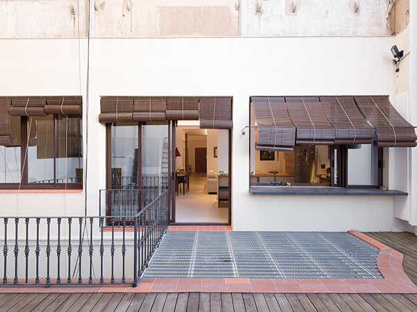 Outdoor patio with metal railing, wooden deck, and large windows with bamboo shades, leading into a modern interior.