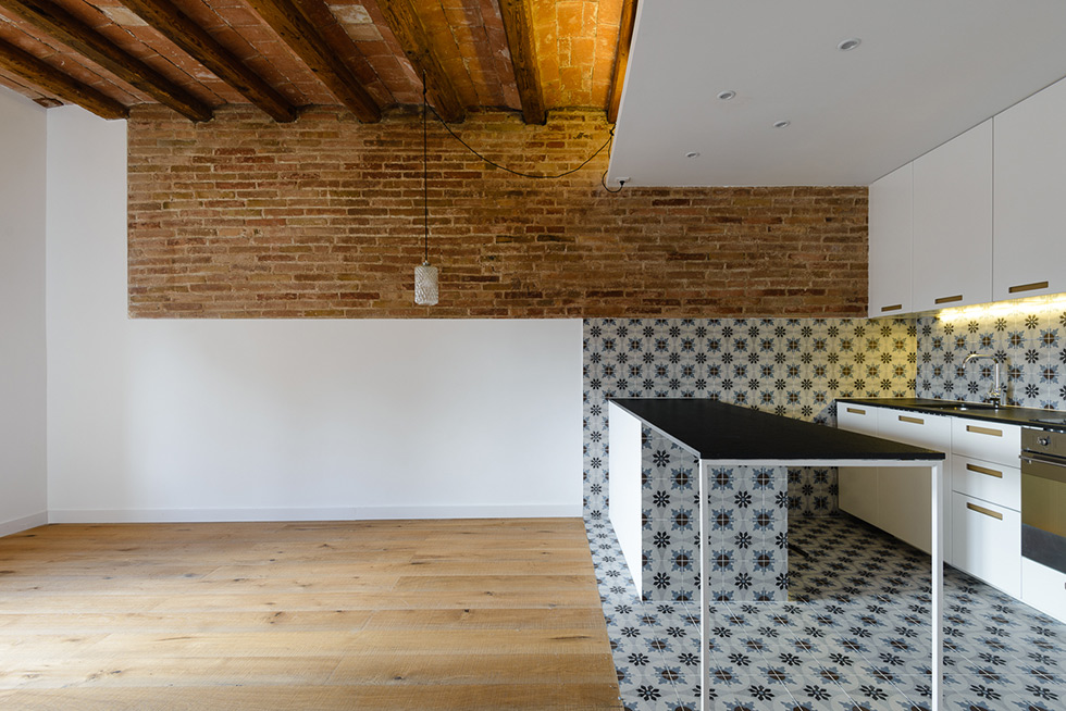 Modern open kitchen with a black work table, patterned tiles, exposed brick wall, wooden beams, and white cabinets.