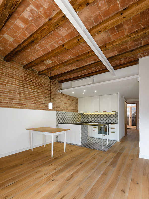 Open-plan kitchen with patterned tiles, exposed brick wall, wooden beams, and white cabinets.