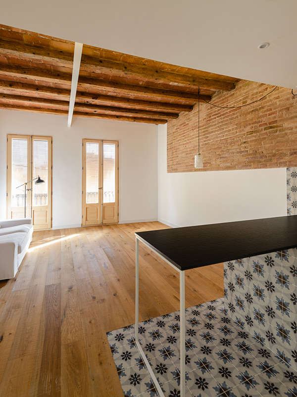 Open-plan kitchen and living area with patterned tiles, exposed brick wall, wooden beams, and two large sunlit balconies on the background.