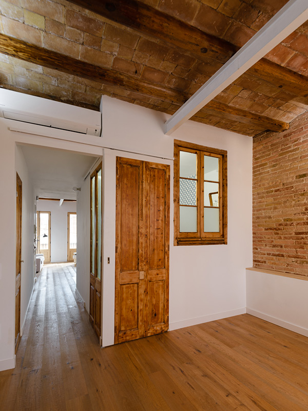 Hallway communicating two living areas, with wooden doors and windows, exposed brick walls, and wooden beams.