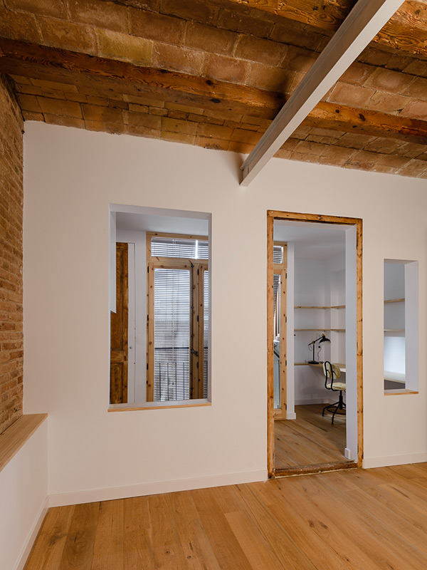 Interior with exposed brick ceiling, white walls, wooden frames, and a view into a workspace with wooden floors and windows.