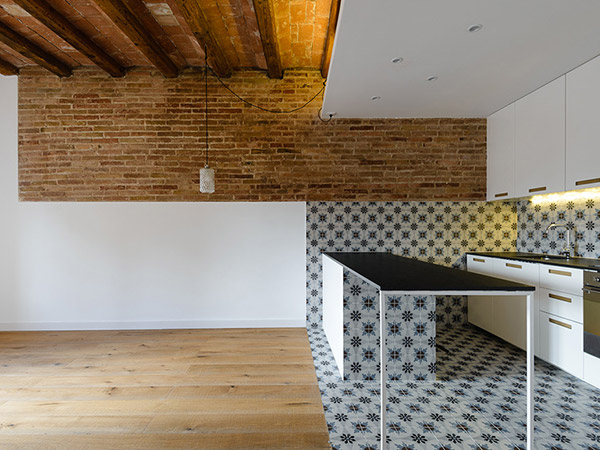 Modern open kitchen with a black work table, patterned tiles, exposed brick wall, wooden beams, and white cabinets.