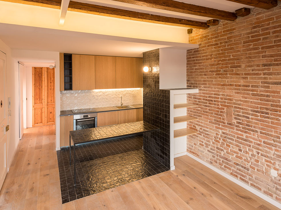 Modern open kitchen with exposed brick walls, light wooden floors, and black tiled counter with a black tiled work table under wooden beam ceiling.