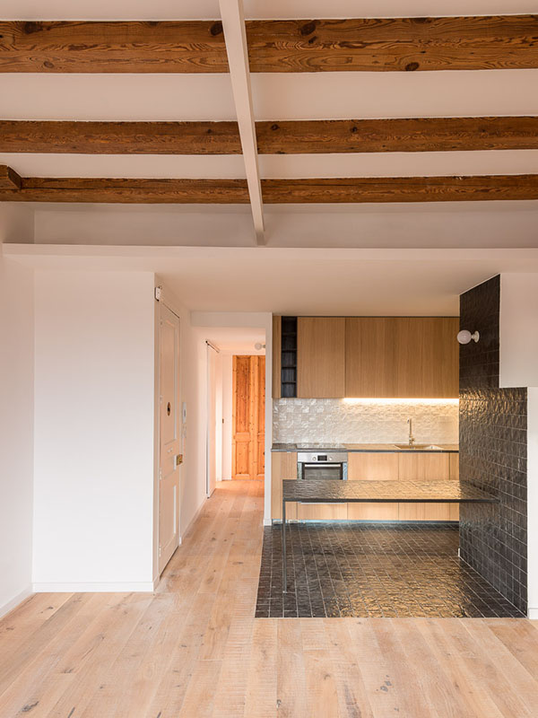 Open kitchen with wooden cabinets, black tiled countertop, and wooden beams on the ceiling.