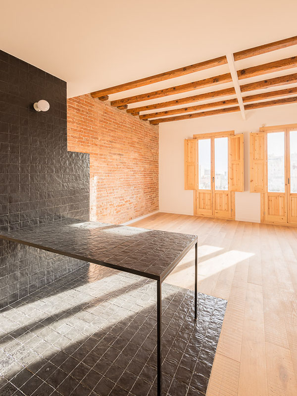 Open-plan living area with black tiled countertop, exposed brick wall, wooden ceiling beams, and natural sunlight coming through two large balconies with wooden shutters.