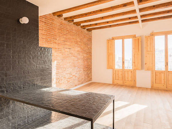 Open-plan living area with black tiled countertop, exposed brick wall, wooden ceiling beams, and natural sunlight coming through two large balconies with wooden shutters.