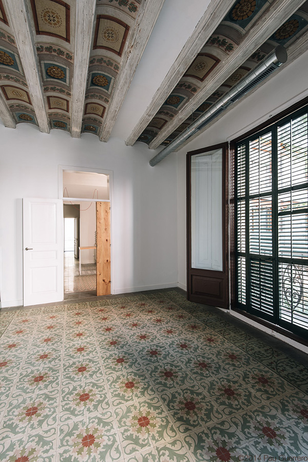 Room with ornate ceiling beams and patterned floor tiles, featuring a partially open wooden door and shuttered window.