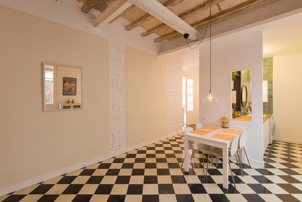 Modern dining area with checkered floor, exposed beams, and white brick accents, featuring a small dining table and pendant light. A square mirror on the wall reflects a door opening into a patio