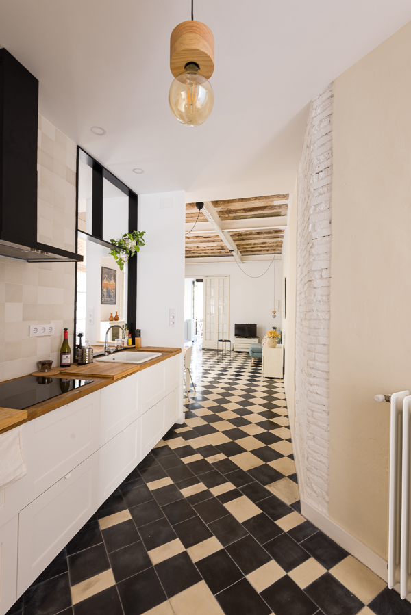 Modern kitchen with checkered floor, white cabinetry, wooden countertops, and a stylish pendant light. In the background, a large living area with checkered floor and wodden beams.