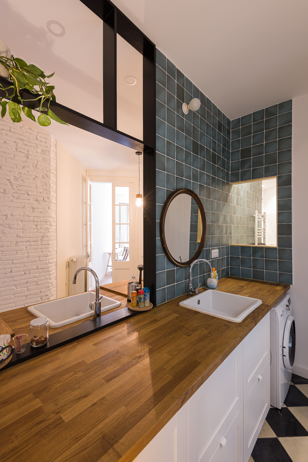 Modern kitchen with wooden countertops, white cabinetry, and teal tiled walls, featuring a round mirror and two facing sinks.