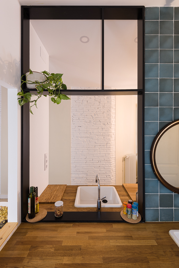 Kitchen sink area with a wooden countertop, slate blue tiles, and a black-framed window opening onto the facing sink area.