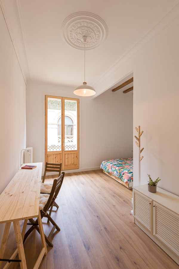 Bright room with wooden floors, a studio desk, and a view into a colorful bed and a balcony.