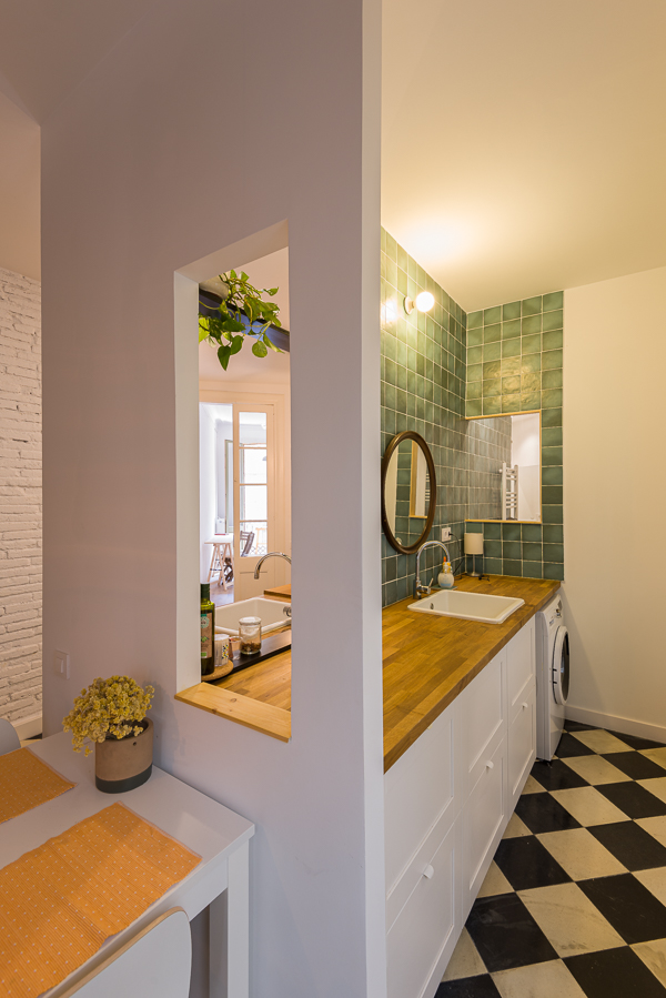 Cozy kitchen with wooden countertops, slate blue tiles, a sink and a washing machine below the countertop. In the foreground, a dining table with yellow mimosa flowers.